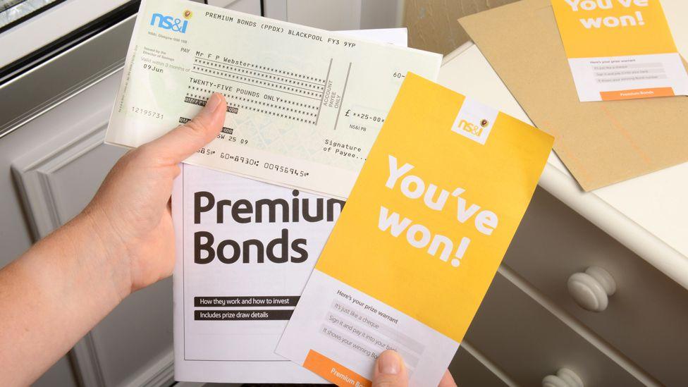 A person's hands holding Premium Bonds literature, including a leaflet saying they have won a prize and a cheque for £25. Part of a white door with a silver letterbox is to the left and a light brown envelope and another NS&I leaflet are on top of a set of drawers to the right