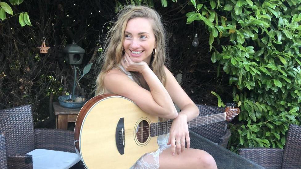 Talia Dean sat in a garden smiling with a guitar on her lap and arm draped over it, looking to the side