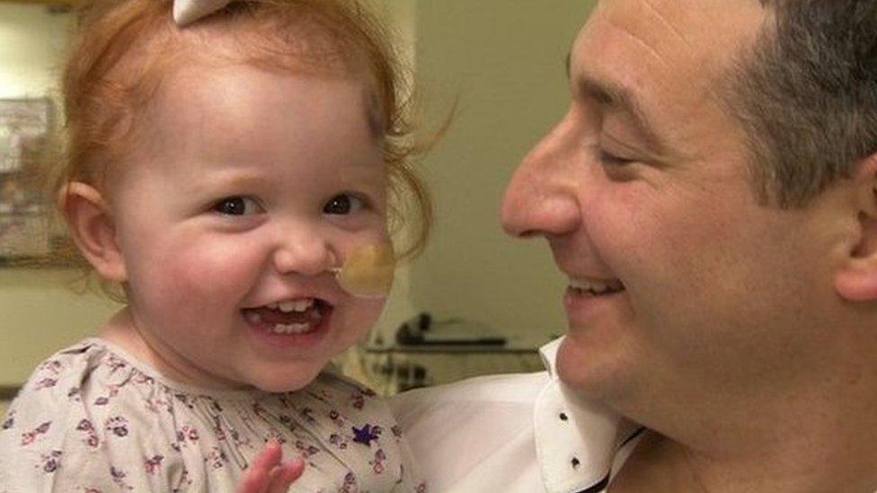 A toddler with ginger hair smiles at the camera, she has a tube taped to her cheek, going into her right nostril. She wears a white top with purple flowers on it. She is held by a man in a white shirt who smiles at her.