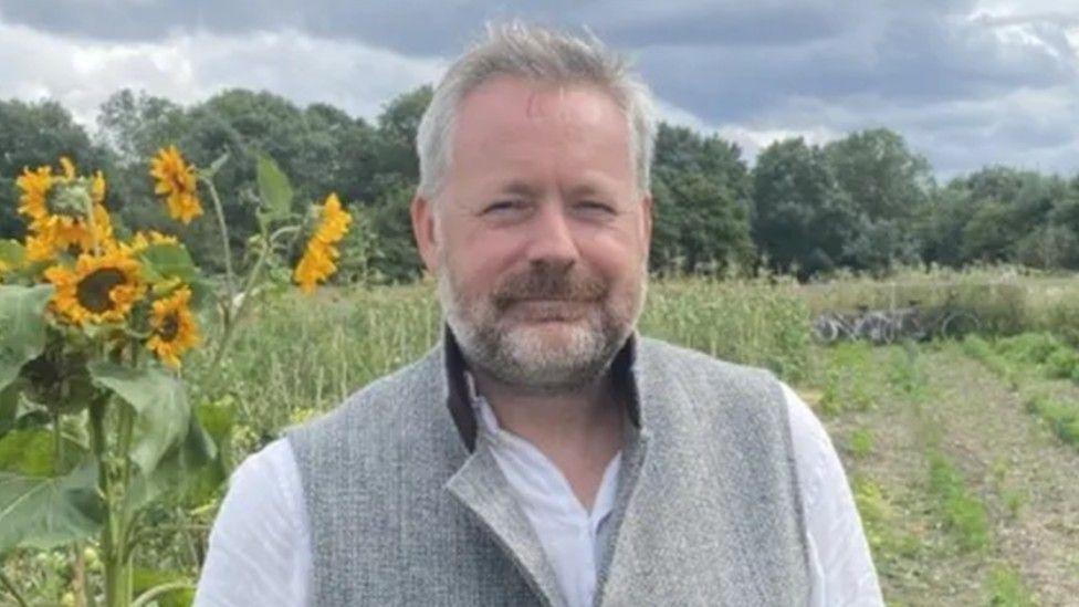 Gavin Shelton, a man who is standing in a field in front of some yellow sunflowers. He is wearing a white linen shirt and grey gilet.