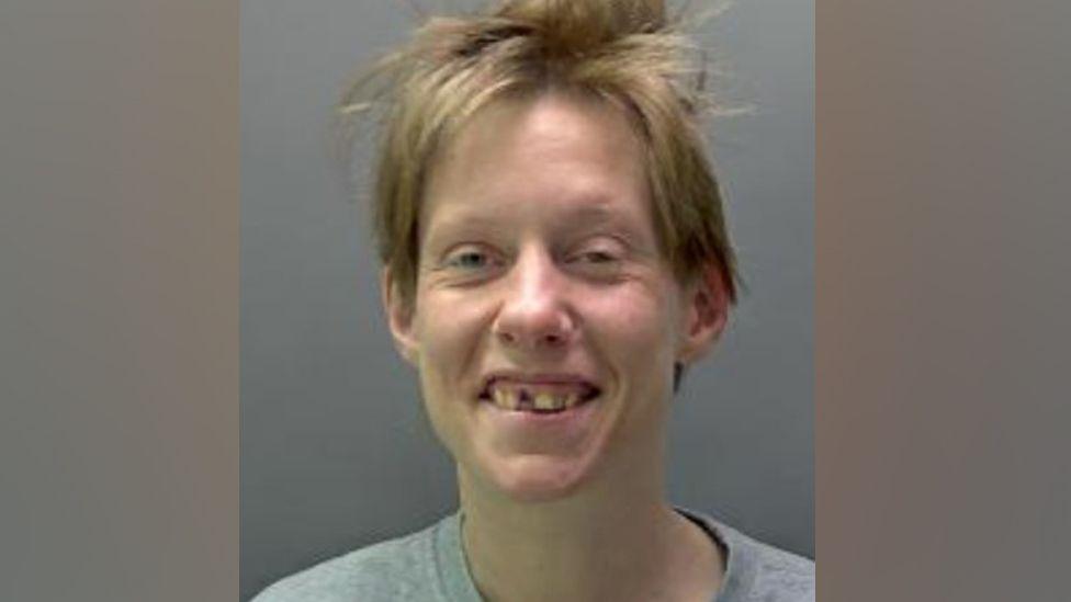 Erin McNeilly is seen in a police custody image the woman with short brown hair, smiling. She is wearing a grey round-necked T-shirt and one of her front teeth is missing.