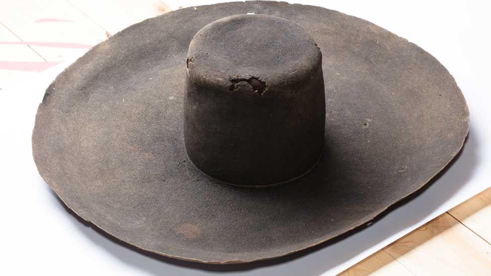 A very broad brimmed black hat resting on a table, dating to the 17th Century. It has become rusty coloured over the centuries and its crown is missing some small pieces.