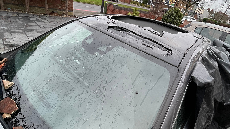 A black car with badly damaged sunroof and cracked windscreen. Black plastic is being used to keep the elements out