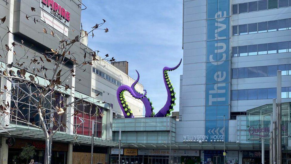 Purple tentacles with green suckers coming out vertically from a shopping centre roof