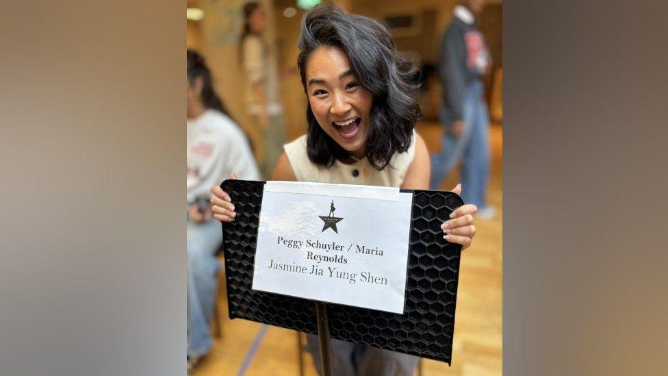 Jasmine has shoulder-length black hair. She is smiling and holding a board which has a white piece of paper stuck to the back. It reads "Peggy Schuyler / Maria Reynolds. Jasmine Jia Yung Shen"