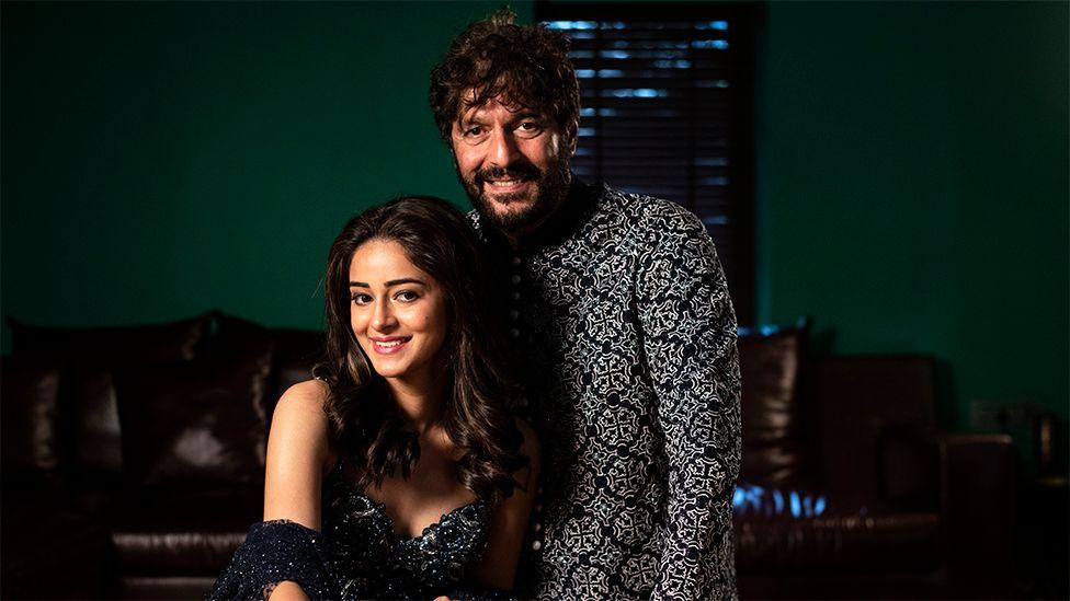 Ananya Panday sitting in her home, with her father Chunky Panday standing next to her. They are wearing sparkling traditional Indian outfits and smiling. Behind them in the room is a brown leather sofa and green coloured walls.