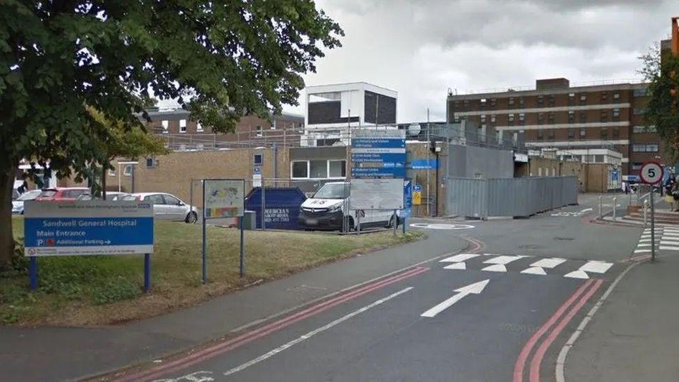 A Google street view of the outside of Sandwell General Hospital. A road is leading to the entrance of the hospital with cars parked on the left hand side.