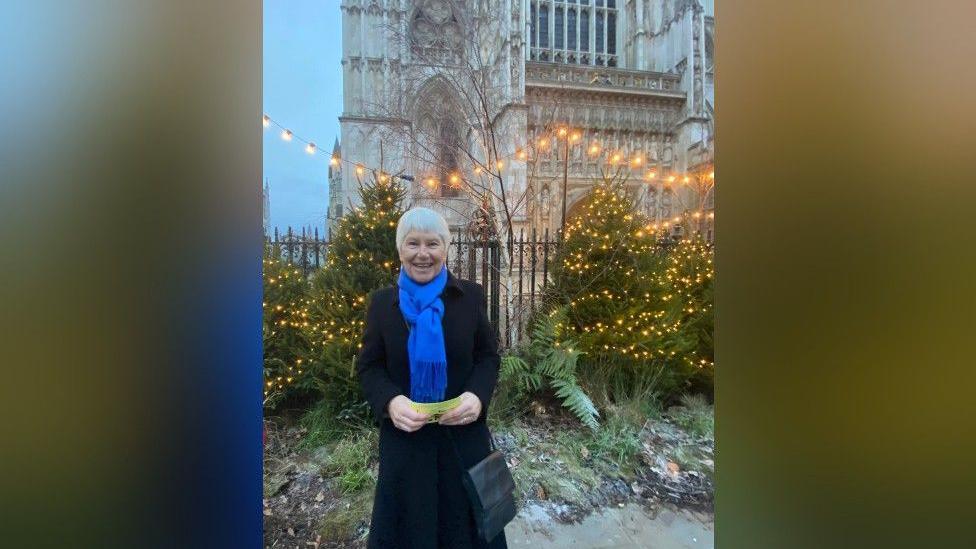 Louise is standing next to an iron fence, with Westminster Abbey in the background. There is a string of yellow lights above the fence. Louise is in a long black coat with a blue scarf. She has white hair and is smiling at the camera. She is holding a yellow piece of paper in her hands.