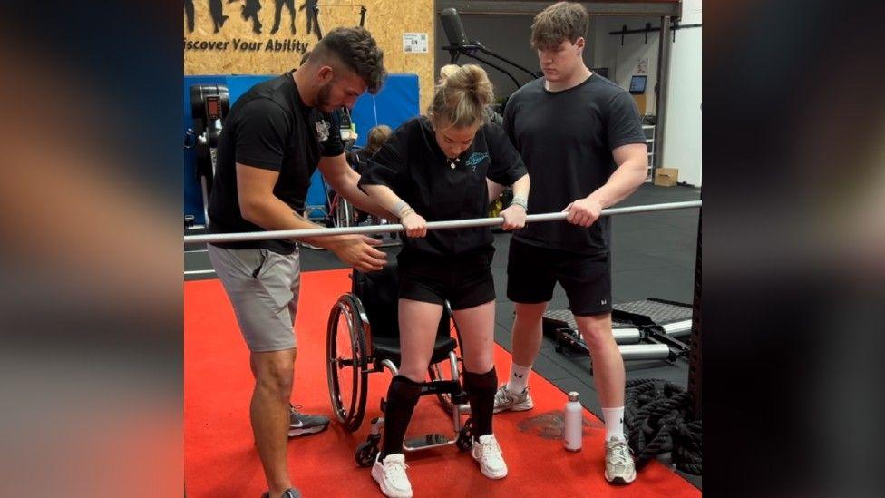 Emma Daniels in a gym wearing all black sports gear. She is standing with the help of two men either side of her and she is holding a metal rod. The gym has red flooring.