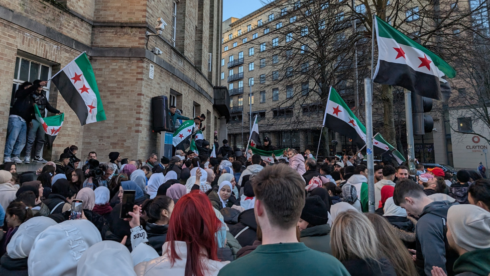 Large crowd of people waving Syrian flags outside Broadcasting House in Belfast, some men have climbed the building and have a large speaker... Clayton hotel and BBC building visible 