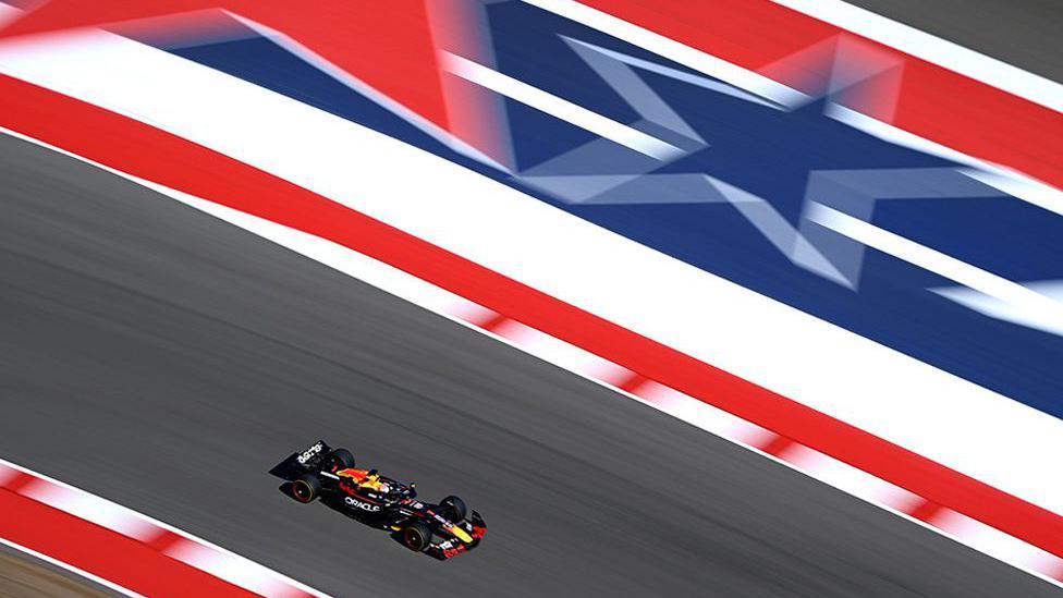 A Formula 1 car with Red Bull and Oracle branding speeds along a track featuring a large star design in red, white, and blue, resembling the Texas flag.