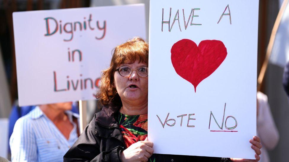 A woman holds up a sign urging MSPs to vote against the assisted dying bill. She has red hair and glasses, and is wearing a dark jacket over a red and green patterned top.