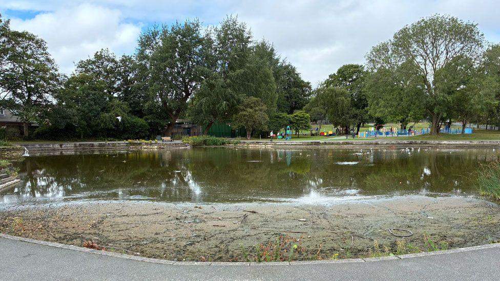A view of the lake showing a large area is dry and there is silt and debris. A children's play area is in the background.
