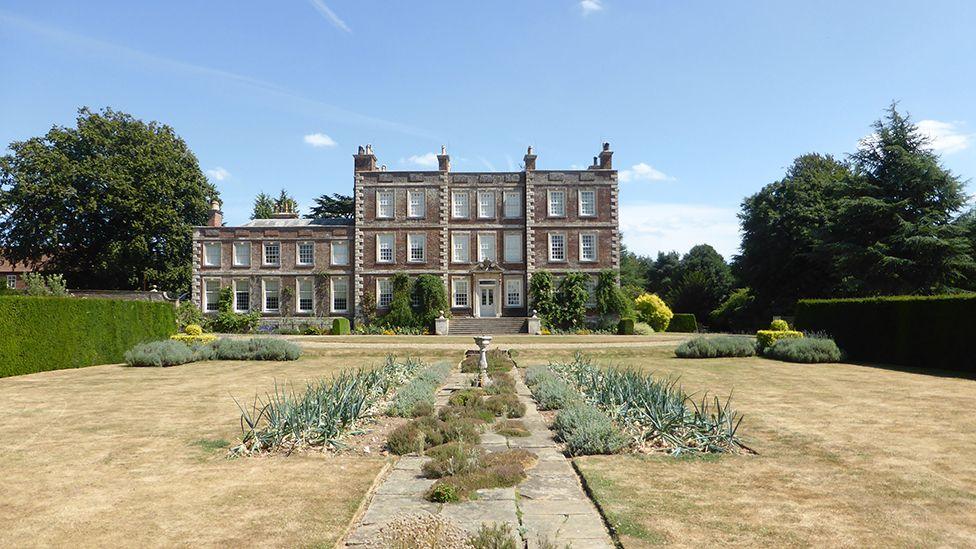 Exterior of Gunby Hall a three-storey brick building with a two-storey annex on the left. It has a large lawn at the front and steps leading up to a white front door.