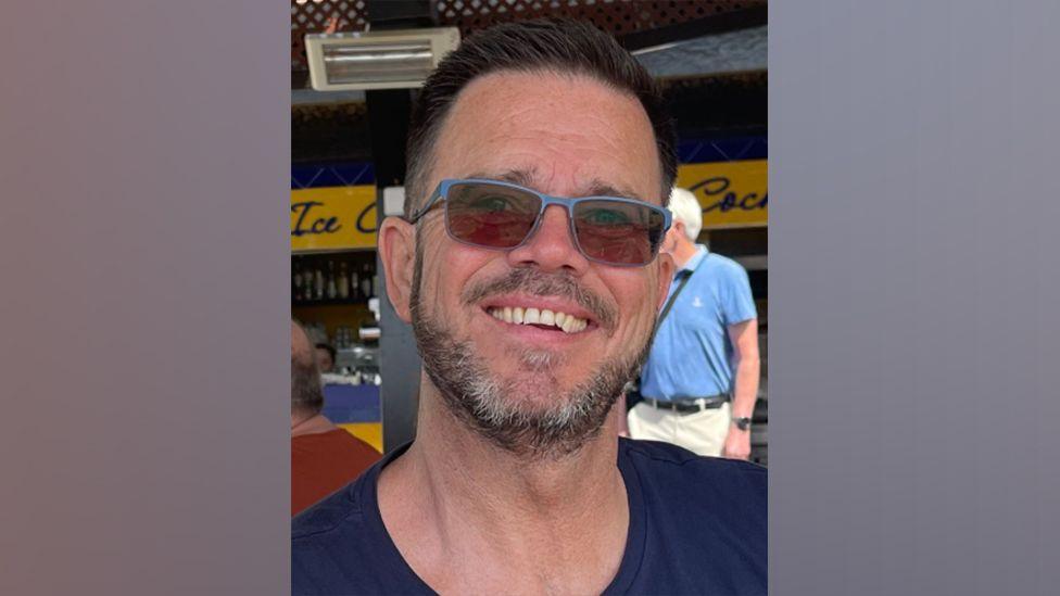 Jonathan Neilan smiling while sat outside under shade on a sunny day outside a restaurant. He is wearing a navy top and sunglasses.