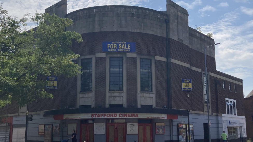 A 1930s cinema building. A sign at the front says "Stafford Cinema" and there are for sale signs on the walls.