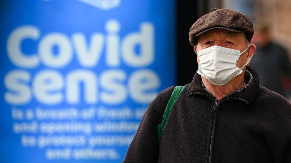 An elderly man wearing a flat cap walks along a street with a face mask on. A blue billboard reading "Covid Sense" can be seen blurred in the background