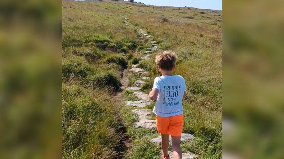 Freddie running uphill away from the camera, wearing a t-shirt and shorts, on a clear summer's day along a narrow path with rocks as stepping stones through a field of heather 