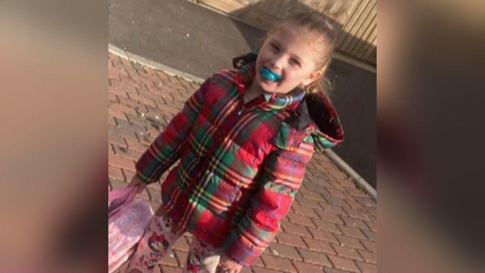A little girl with blond hair. She has a red checked puffer jacket on and a dummy in her mouth. She is holding a pink backpack. 