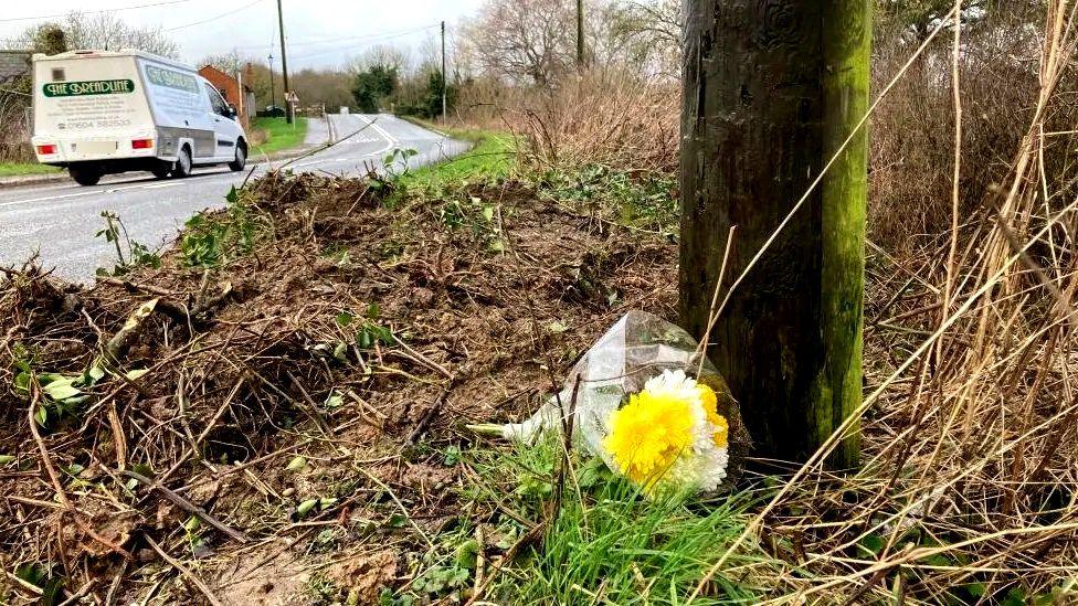 A roadside shrine showing a bouquet of yellow flowers at the base of a wooden post, which is covered in green moss. The mud alongside the post has been churned up by a vehicle. There is a road running past and a white van with green writing is driving away from the camera.