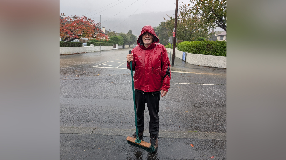 Peter Law. He is standing at the end of a street holding a brush. He is wearing a red raincoat and black trousers.