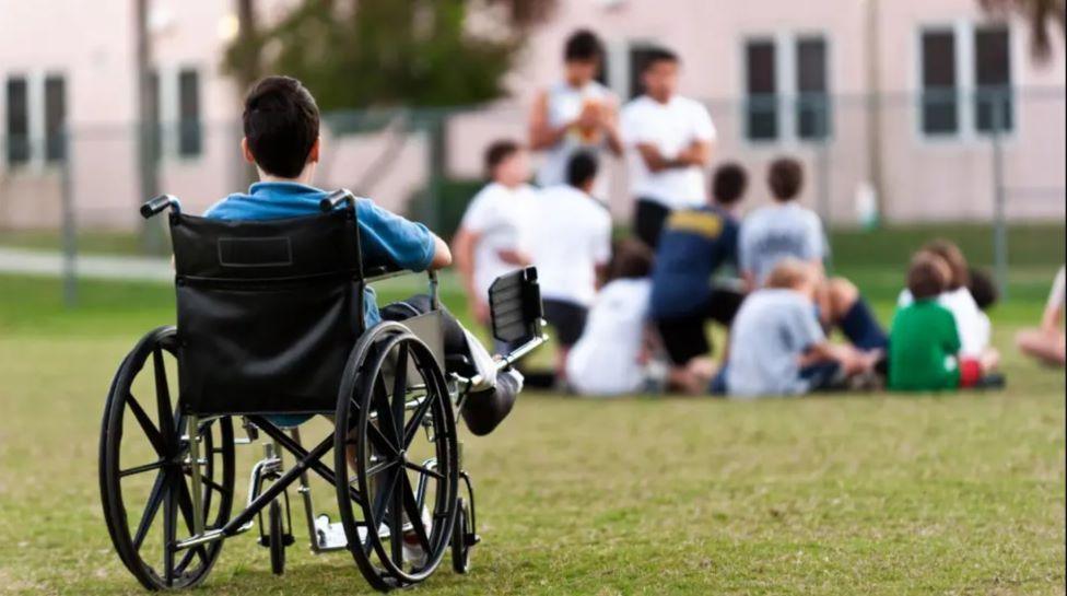 A child who uses a wheelchair has his back to the camera. He is wearing a blue top and is looking towards another group of children.