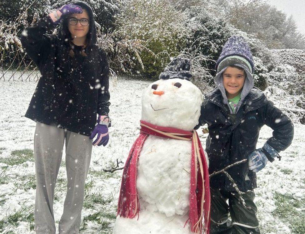 Two children stand either side of a snowman with a carrot nose and scarf