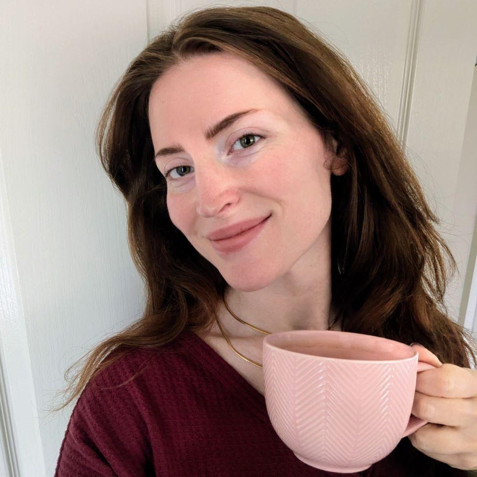A woman with brown hair smiles and looks at the camera, holding up a pale pink mug
