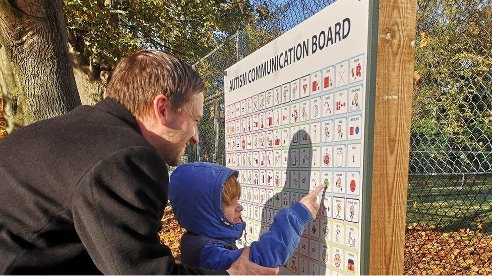 Dan wearing a grey coat looking at the white image board with Joshie - who is wearing a blue hoodie and pointing to the board in a park