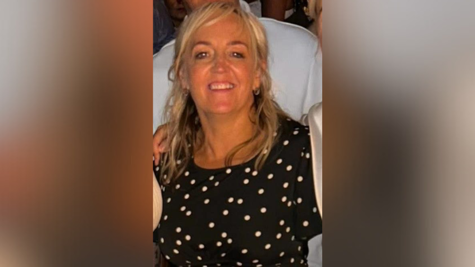 A woman is smiling into the camera. She has long blonde hair. She's wearing a black and white polka dot dress and hoop earrings. Other people are standing close to her and an arm is draped over her shoulders.