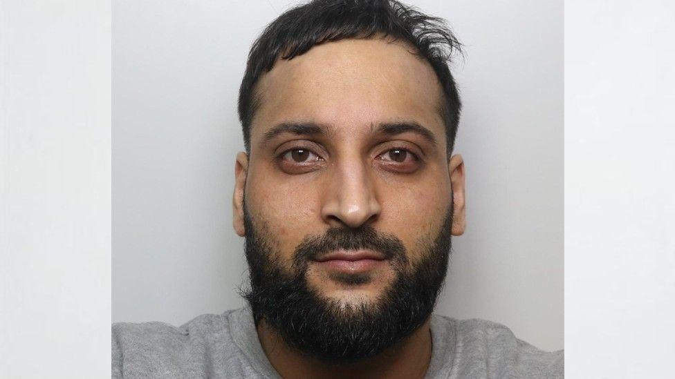 Hussain in a police custody picture. He has black, short hair and a black, trimmed beard. He is wearing a grey jumper 