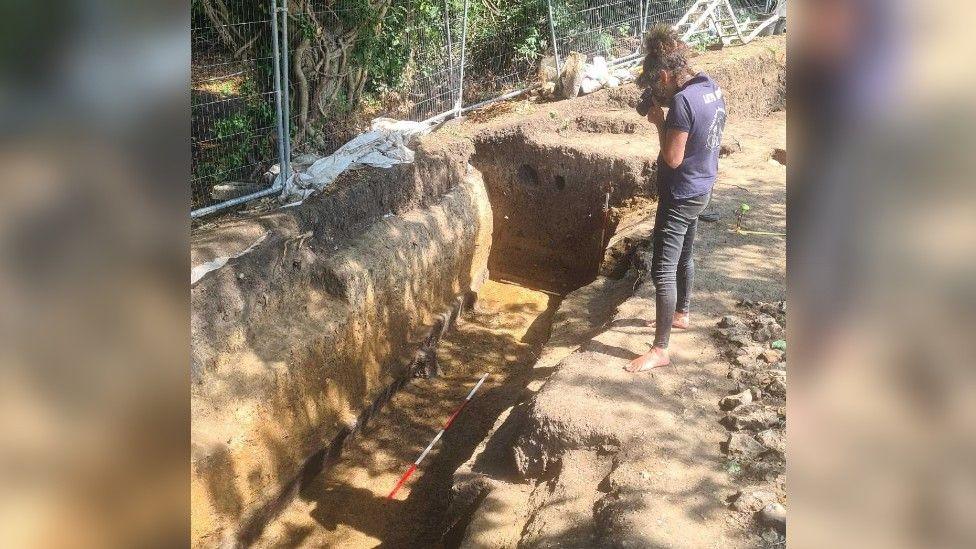 Archaeology expert  wearing black jeans and purple t-shirt taking a photo of the trench