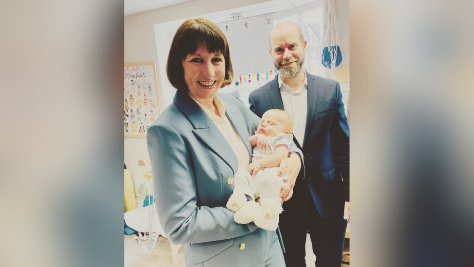 Woman on the left wearing a light blue trouser suit with a white blouse and short brown hair is holding a newborn baby who is wearing a cream baby grow and cream boots. Man with a moustache and beard on the right wearing a navy blue suit with a white shirt.