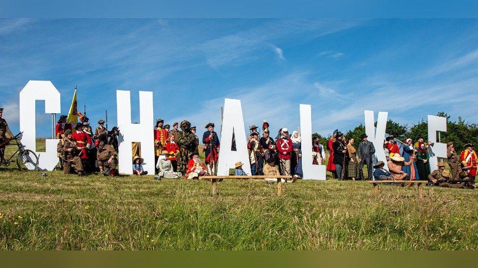 Large white festival letters spelling out Chalke on a grassy hill underneath a blue sky - each are surrounded by re-enactors, all in costume, some red military uniform, others more recent, some in medieval dress too.