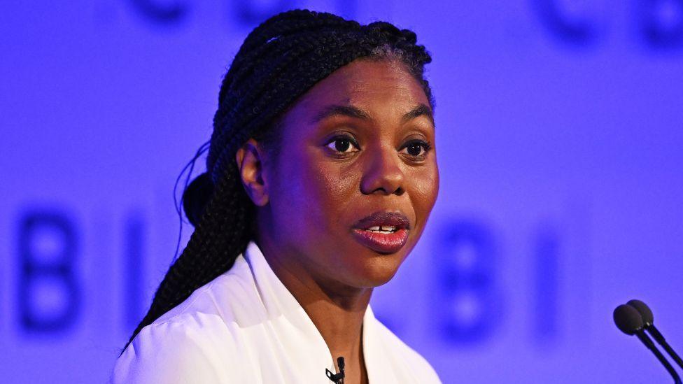Kemi Badenoch, who has long dark hair, speaks into a microphone. She is wearing a white top, with a wall illuminated with blue lights behind her. 