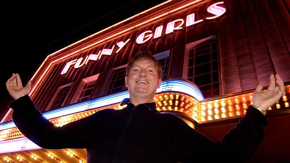 Basil Newby stands under the neon lit Funny Girls venue at night with his arms outstretched