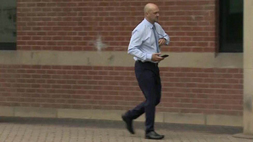 A side-on picture of former PC Neal Murray walking to court for a previous hearing. He is wearing a light blue shirt and tie, dark blue trousers and black shoes. In his right hand he is carrying a mobile phone at waist height.