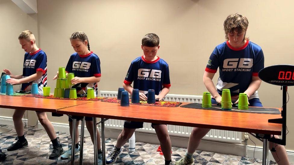 Four young people stand behind a long table indoors, each using plastic cups arranged on mats to practise sport stacking. They wear matching blue shirts with “GB Sport Stacking” printed on the front, and stacks of green and blue cups are visible on the table as they work.