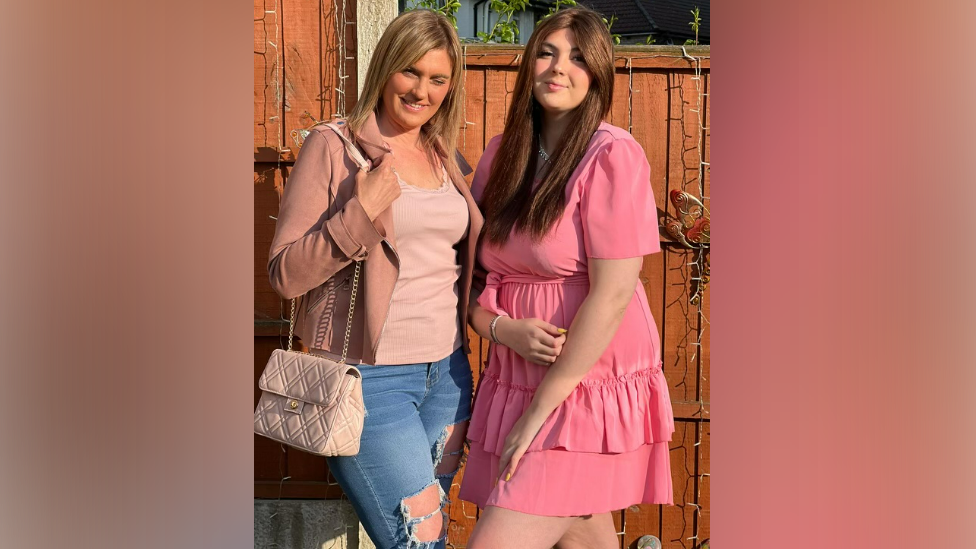 Two women pose in the garden and smile for the camera. Jenny, to the left, is wearing a pink jacket and top with ripped jeans and a handbag, while Leah, to the right, is wearing a pink ruffle dress. 