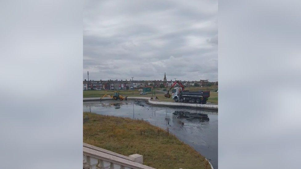 Clean up of Coatham boating lake. A lorry and an excavator are removing items from the nearly empty lake.  There is a row of houses in the background. 