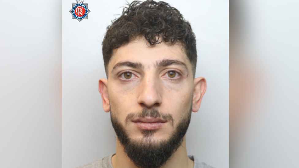 A police mugshot of a man looking at the camera. He is expressionless and has a dark hair, a dark beard and moustache.