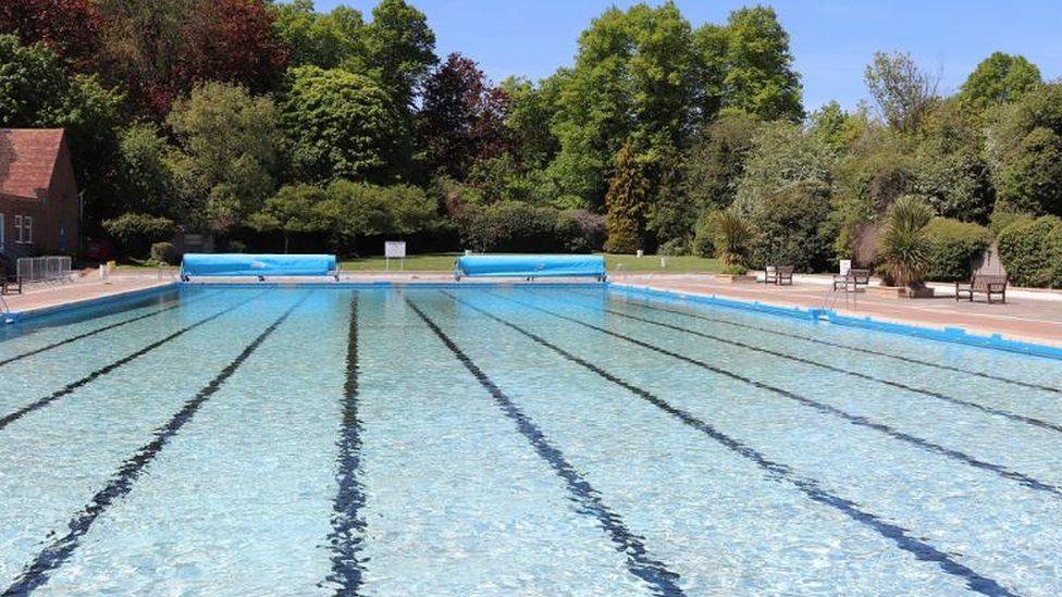Letchworth Lido, showing a large expanse of blue water. There are lane lines marked out on its floor. On the right is a tiled area with benches. At the end of the pool are rolled back blue pool covers, beyond is is grass and trees in full leaf.