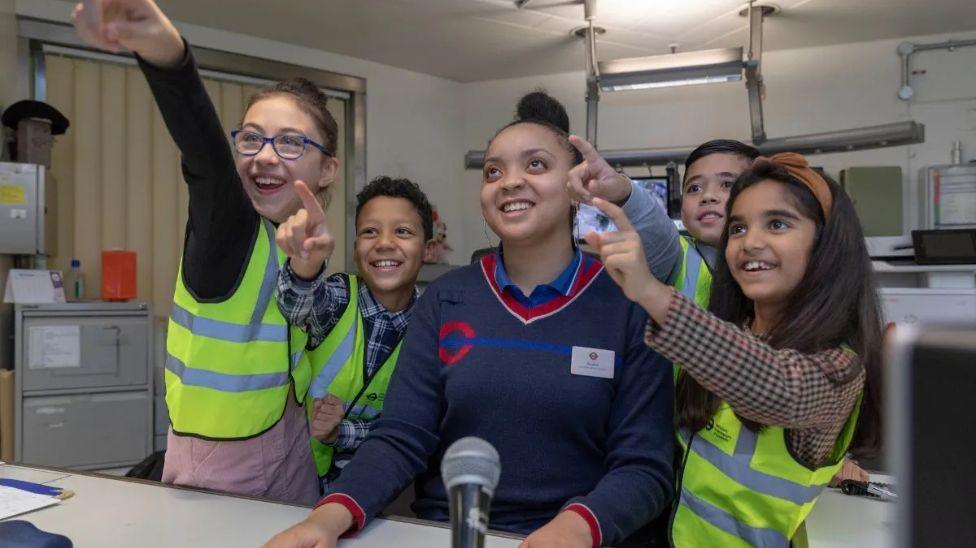 four children with a Transport for London staff member wearing high vis jackets and pointing at something.