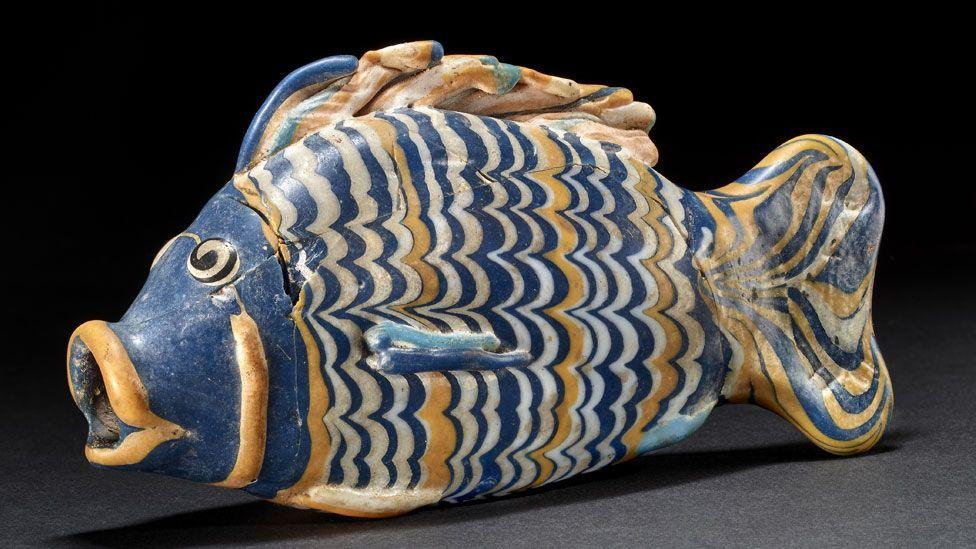 A blue, yellow and white glass vessel in the shape of a fish. Its body, tail and top fin have wavy stripes in blue, yellow and white, its face is blue, its open mouth outlined in yellow. It is resting on a black surface.