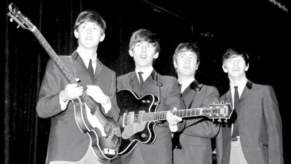 A black and white photograph of all four members of The Beatles holding guitars backstage on their 1963 tour.