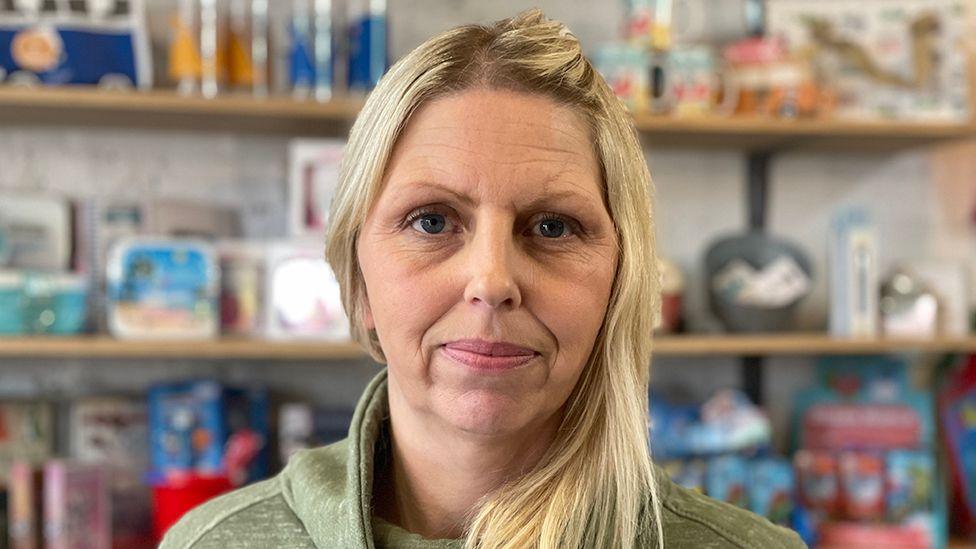 Sophie Kirk with blond hair wearing a green top standing in front of a row of shelves in her shop