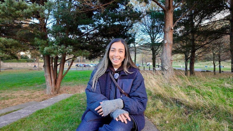 A picture of Devamsha sitting in a park wearing a dark blue coat, surrounded by grass and trees.