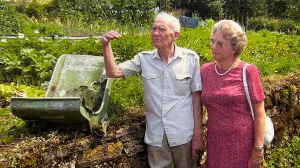 An elderly man stands hand in hand with a woman. To his right is a battered world war 2 plane seat sat on a garden wall