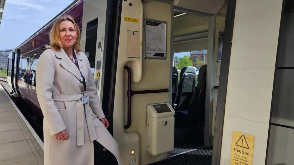 Philippa Cresswell with long blonde hair wearing a long beige coat. She has one leg on the step by the entrance to the coach of a train. She is standing on a train platform.