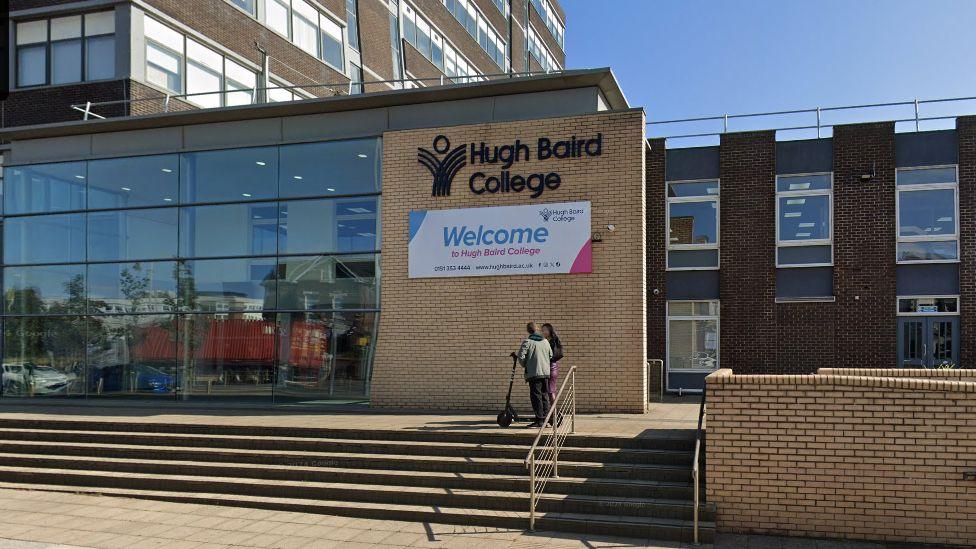 Street view of Hugh Baird College in Bootle, showing wide flagged steps leading up to a large glass entrance with a welcome sign